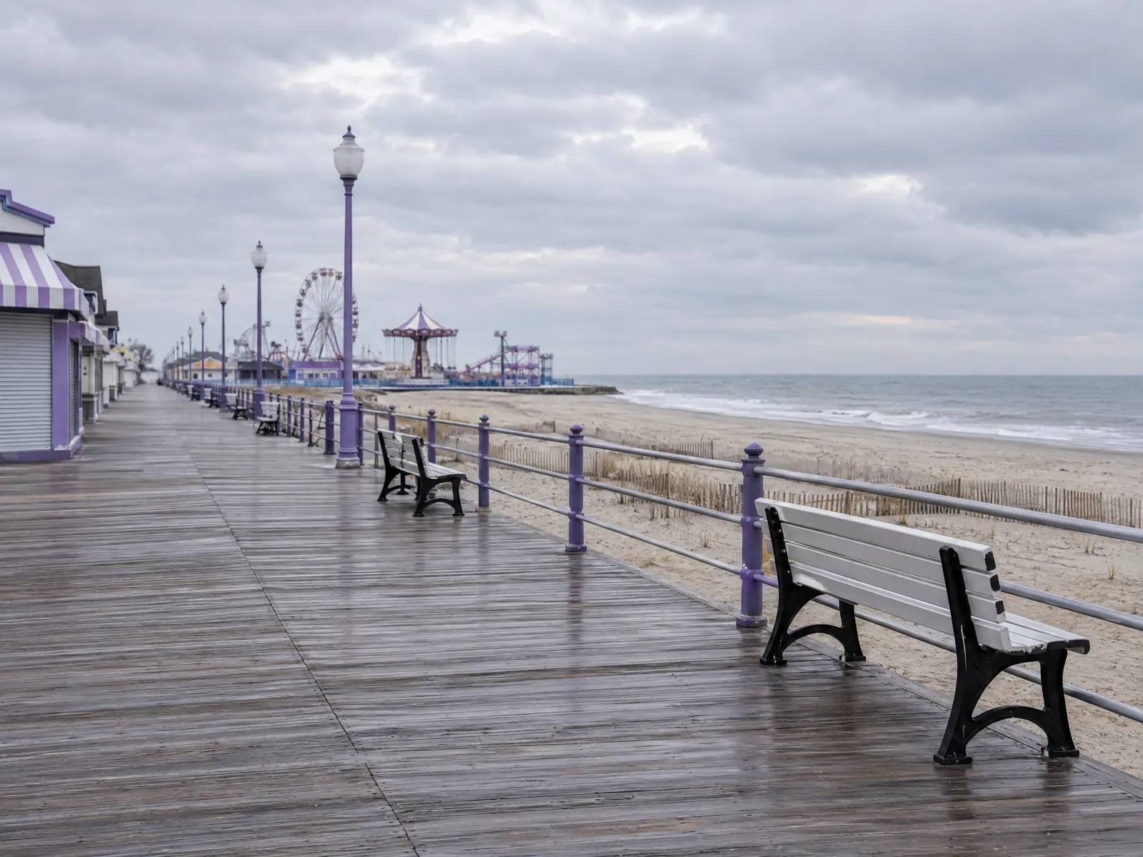 Empty wooden boardwalk with benches beside a winter beach and distant carnival rides